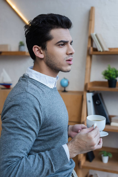 Thoughtful businessman holding saucer and cup of coffee in office - Photo, image