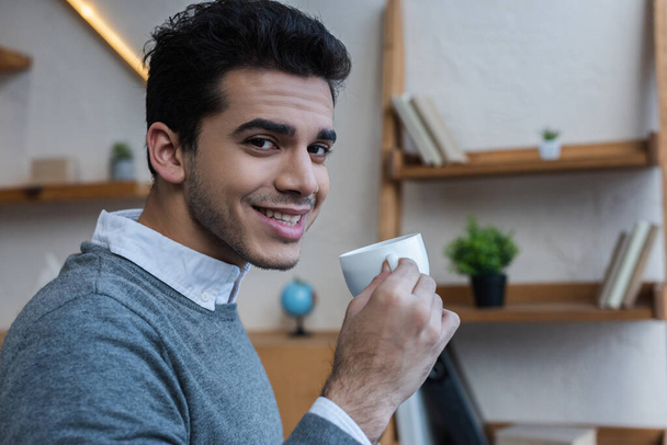 Businessman smiling, looking at camera and holding cup of coffee in office - Valokuva, kuva