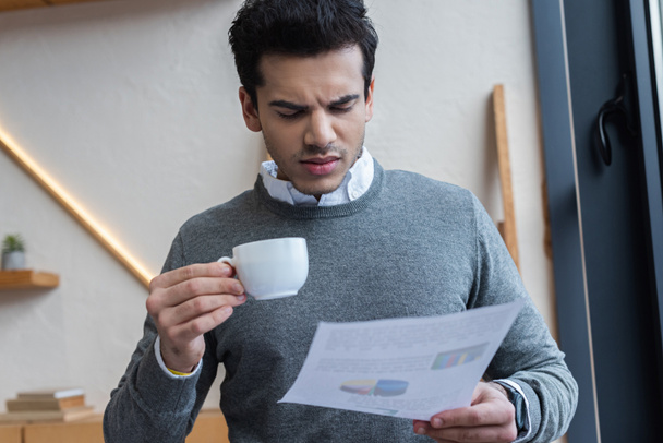 Zamyšlený a ustaraný podnikatel s šálkem kávy při pohledu na papír v kanceláři - Fotografie, Obrázek