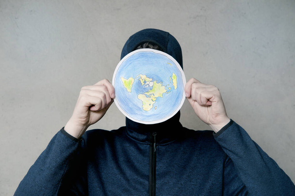 Flat Earther concept. Person who believes that Earth is flat disc. Anonymous hooded Man holding flat Earth model in front of face, copy space. Isolated on gray background, studio shot. - Photo, Image