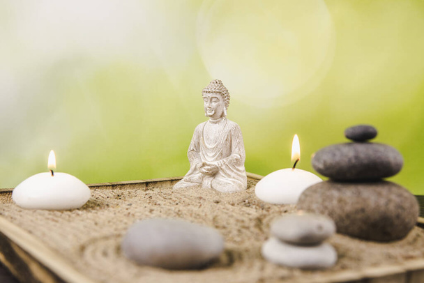 Miniatur-Schreibtisch-Zen-Sandkasten mit Buddha-Figur in Lotus-Position, gestapelte Zen-Meeressteine, Wellness-Kerzen, die vor grünem Bokeh-Studiohintergrund brennen, Kopierraum. - Foto, Bild