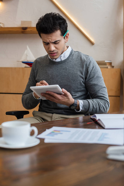 Selective focus of worried businessman holding digital tablet at table in office - Photo, image