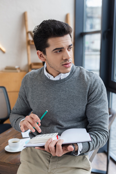Задумчивый бизнесмен с блокнотом, карандашом и калькулятором смотрит в сторону офиса
 - Фото, изображение