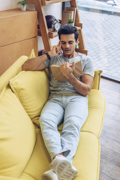 Високий кут зору людини з книгою розмовляє на смартфоні і лежить на дивані у вітальні
 - Фото, зображення