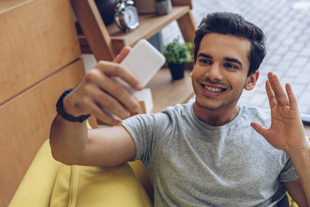 Selective focus of man waving hand, smiling and taking selfie with smartphone on sofa - Foto, afbeelding