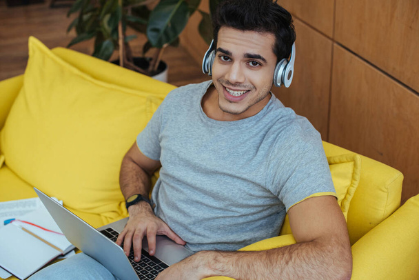 High angle view of freelancer in headphones with laptop smiling and looking at camera on sofa - Foto, Imagen