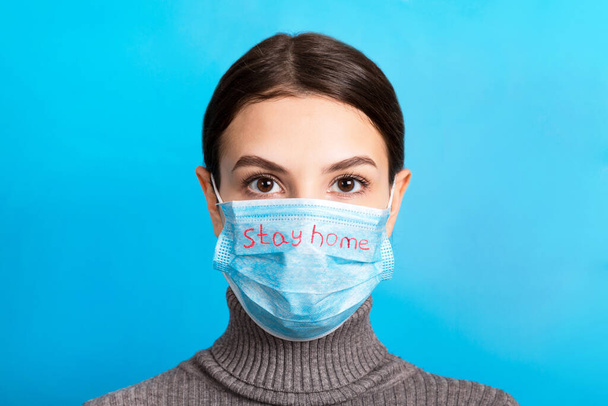 Portrait of pretty woman wearing medical mask with Stay home text at blue background. Coronavirus concept. Respiratory self isolation protection. - Photo, Image