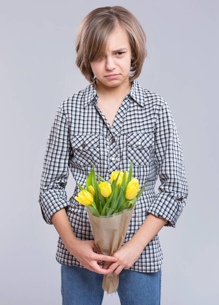 Beautiful sad girl with bunch of flowers on gray background. Child with bouquet of yellow tulips as a gift. Happy mothers, Birthday or Valentines day. - Photo, image