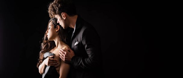 panoramic shot of handsome man in suit hugging sensual nude woman in black cloth isolated on black - Photo, Image