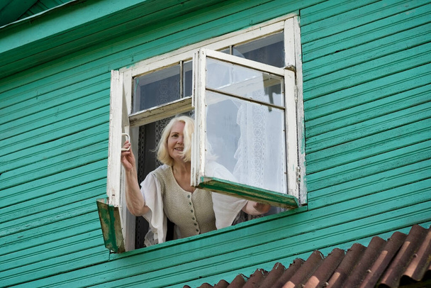 Ανώτερος καυκάσιος γιαγιά γυναίκα στέκεται κοντά στο παράθυρο κοιτάζοντας εξωτερική σε πράσινο ξύλινο σπίτι στην απομόνωση ή καραντίνα - Φωτογραφία, εικόνα
