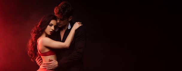 panoramisch schot van vriendje in pak knuffelen sexy vriendin in rood lingerie op zwart met rood licht - Foto, afbeelding