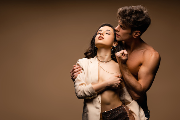 passionate shirtless boyfriend holding necklace of seductive half naked girlfriend in beige jacket on beige - Valokuva, kuva
