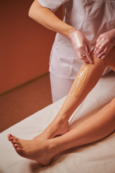 Waxing. The specialist removes hair from the beautiful legs of a young girl using wax. The girl lies on the sofa in the spa and she is doing depilation. - Valokuva, kuva