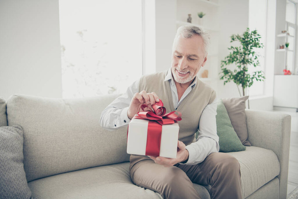 Portrait of cute old man with stubble bristle holding package open wearing trousers shirt sitting divan room - Foto, afbeelding