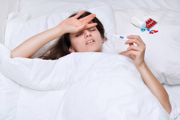 Άρρωστη νεαρή γυναίκα με θερμόμετρο σε λευκό κρεβάτι.Νιώθει την ασθένεια - Φωτογραφία, εικόνα