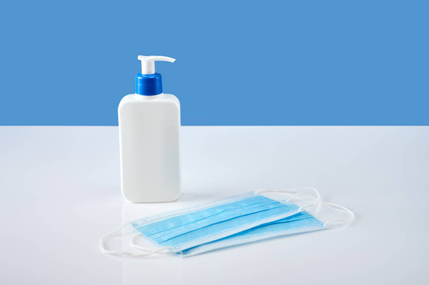 Biała butelka antyseptyczna z dozownikiem i dwie niebieskie maski na białym stole akrylowym, niebieskie tło - Zdjęcie, obraz