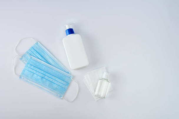 Biała butelka antyseptyczna z dozownikiem obok środka do dezynfekcji rąk, chusteczki antyseptyczne i dwie niebieskie maski medyczne na białym stole akrylowym, widok z góry - Zdjęcie, obraz