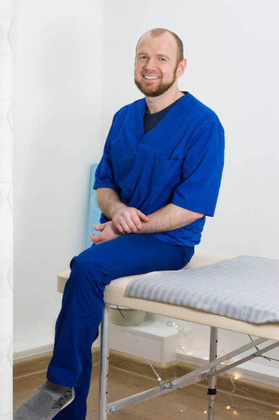 Portrait of a young male doctor. Man in blue uniform of doctor sits on massage table - Photo, Image