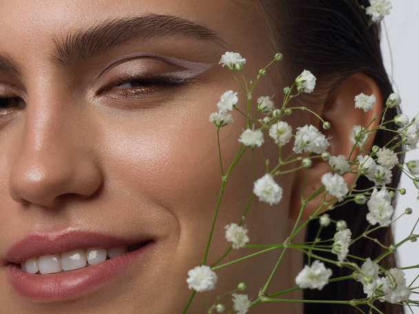 Close up portrait of a face with beautiful skin and nude makeup, eyeliner and long eyelashes, puffy natural lips and white flowers. Fashion photography. Cosmetology and spa. Freshness of spring - Foto, afbeelding
