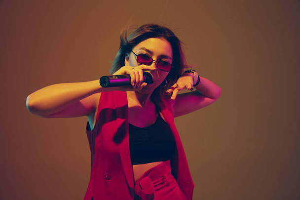 Young caucasian female musician, performer singing, dancing in neon light on gradient background - 写真・画像