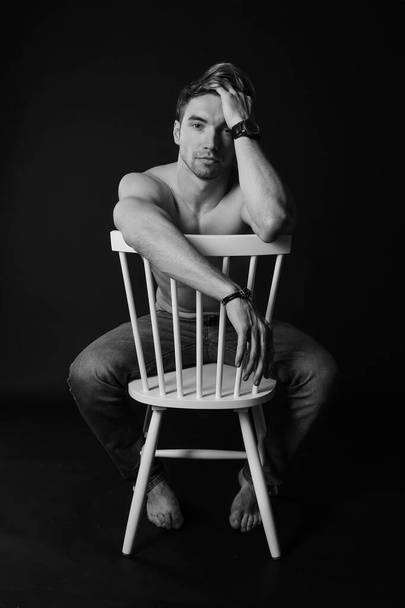 Muskularny i sprawny młody kulturysta fitness męski model pozowanie na krześle. Czarno-białe zdjęcie. - Zdjęcie, obraz