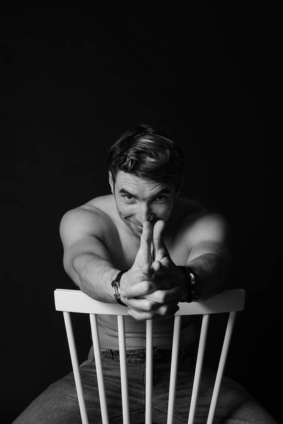 Muscular and fit young bodybuilder fitness male model posing on chair. Black and white photo. - Photo, Image
