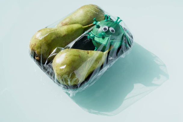 Vista conceptual de una bandeja negra con tres peras naturales y un virus verde juguete que ha contagiado la fruta fresca que viaja en el mismo recipiente envuelto con plástico transparente
. - Foto, imagen