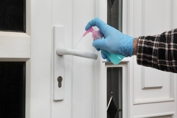 Huna hand in protective glove spraying antibacterial or cleaning solution from sprayer bottle to handle of house door - Foto, imagen