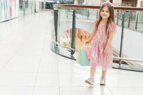 Портрет маленької щасливої дівчинки в торговому центрі. Усміхнена дівчина сміється в рожевій сукні з різнокольоровими сумками в руках ходить навколо торгового центру, дивлячись на вікна магазину
. - Фото, зображення