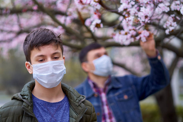 Vater und Sohn mit Gesichtsmaske sind in der Stadt draußen, blühende Bäume, Frühlingszeit, Blütezeit - Allergiekonzept und Gesundheitsschutz vor staubiger Luft - Foto, Bild