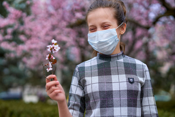 девушка в маске для лица находится в городе под открытым небом, цветущие деревья, весенний сезон, время цветения - концепция аллергии и защиты здоровья от пыльного воздуха
 - Фото, изображение