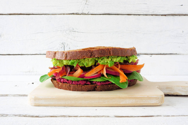Gesundes Superfood-Sandwich mit Avocado, Rübenhumus, Spinat-Radieschen und Karotten. Seitenansicht auf einem Holzbrett vor weißem Holzhintergrund. - Foto, Bild
