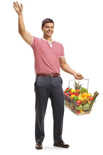 Full length portrait of a young man waving and holding a shopping basket with fruits and vegetables isolated on white background - Foto, immagini