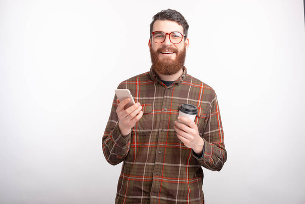 cheerful young man with beard holding smartphone and cup of coffee to go - Foto, Imagen
