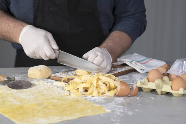 Close-up van een mannelijke chef-kok die een rol zelfgemaakte pasta in noddles snijdt op een snijplank met eieren en eierschalen op een onscherpe achtergrond. Koken en traditioneel voedselconcept. - Foto, afbeelding