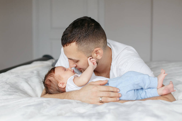Λευκός πατέρας ξαπλωμένος με νεογέννητο μωρό στην κρεβατοκάμαρα. Άντρας γονιός αγκαλιάζει φιλιά αγκαλιάζει παιδί γιο κόρη. Αυθεντικό τρόπο ζωής ειλικρινής στιγμή. Περήφανος νεαρός μπαμπάς. Ημέρα οικογενειακών πατέρων. - Φωτογραφία, εικόνα