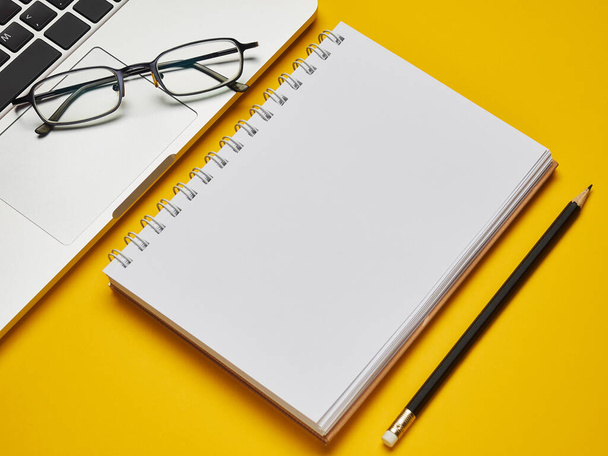 Workplace - laptop, notebook, glasses and pencil for remote work on a yellow background table. Freelance desktop for home or office. Background with copy space. - Photo, image