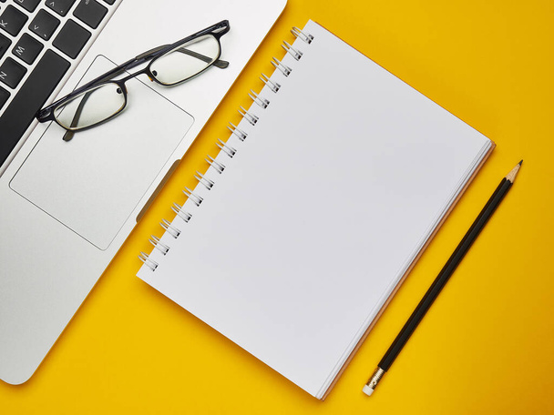 Workplace - laptop, notebook, glasses and pencil for remote work on a yellow background table. Freelance desktop for home or office. Background with copy space. - 写真・画像