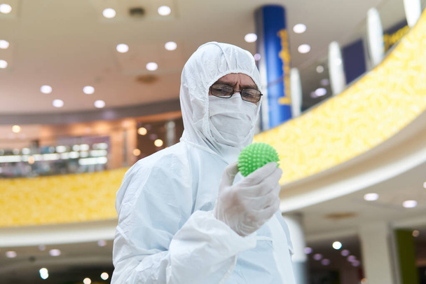 людина в укритті одноразовий анти-епідемічний антибактеріальний ізоляційний костюм підозріло тримає в руці гумовий м'яч, що нагадує макрофото вірусу, на тлі якогось внутрішнього простору
 - Фото, зображення