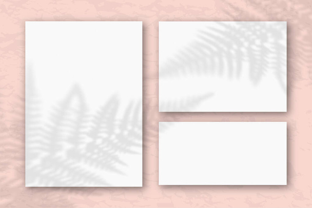 Diversi fogli orizzontali e verticali di carta strutturata bianca contro una parete rosa. Il Mockup si sovrappone alle ombre delle piante. La luce naturale getta ombre dalle foglie di felce. Posa piatta, vista dall'alto
. - Foto, immagini