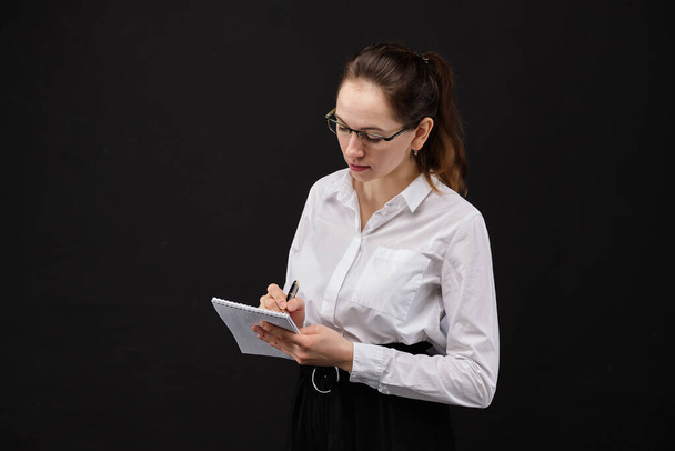 Girl in a white shirt in glasses makes an entry in a notebook on a black background. Close up. - Photo, Image