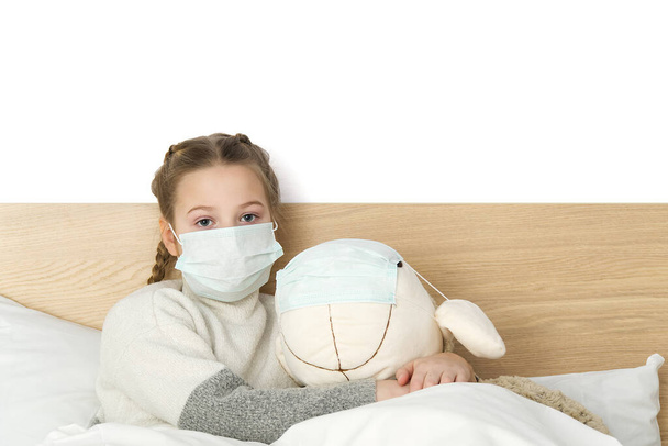Uma criança triste doente em uma máscara com uma temperatura e uma dor de cabeça está na cama. Gripe constipações vírus da doença bacterium. menina com brinquedo no quarto de cama usar máscara de proteção, proteger contra o conceito Coronavirus
 - Foto, Imagem