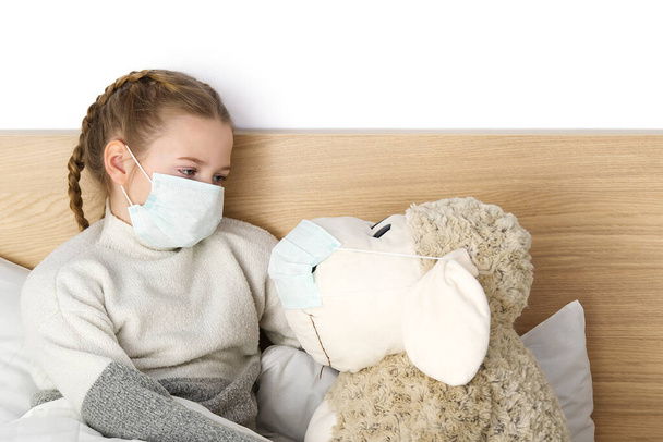 Nemocné smutné dítě v masce s teplotou a bolestí hlavy leží v posteli. Bakterie viru chřipky. dívka s hračkou v ložnici nosit ochrannou masku, chránit proti coronavirus koncept - Fotografie, Obrázek