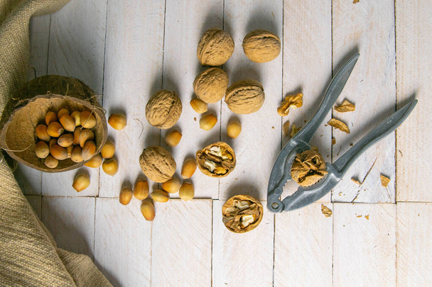 Walnuts in a wooden box next to a hazelnut in a coconut shell, nuts scattered on a white wooden table on which the nut cracker also stands. - Foto, Bild