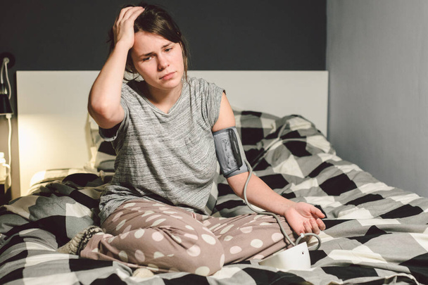 Das Thema Bluthochdruck ist Bluthochdruck. 22-jährige junge kaukasische Frau benutzt ein automatisches Tonometer, um den Druck zu Hause im Schlafzimmer auf dem Bett in Heimkleidung am Abend zu überprüfen. - Foto, Bild