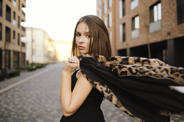 Gorgeous girl walks about the city. Brunette with long hair in a black dress and a leopard cape. Girl, fashion photo, street fashion. Fashionable girl walks in the city of Europe, travels. - Photo, Image