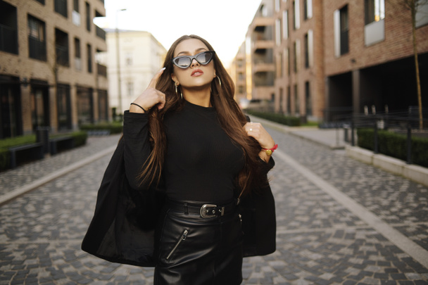 Gorgeous girl walks about the city. Brunette with long hair in a black dress and leather. Girl, fashion photo, street fashion. The model walks around the city of Europe, travels. Girl in fashionable glasses. - Zdjęcie, obraz