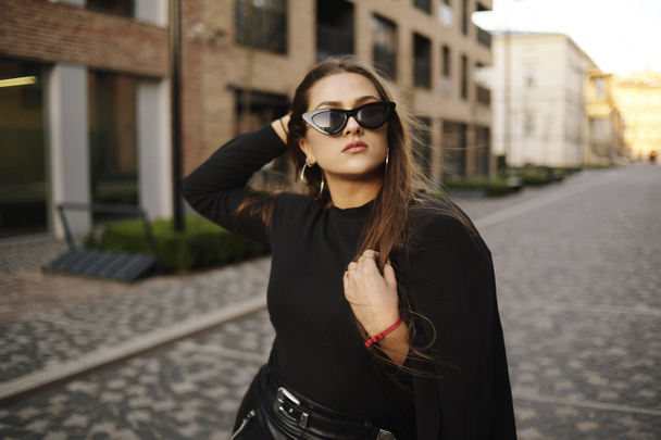Gorgeous girl walks about the city. Brunette with long hair in a black dress and leather. Girl, fashion photo, street fashion. The model walks around the city of Europe, travels. Girl in fashionable glasses. - Photo, image