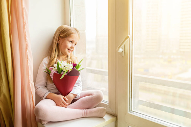 School kid in self isolation sitting near window with bouquet of flowers. Girl stay at home during covid-19 lock down, self isolation, stay home concept. - Фото, изображение
