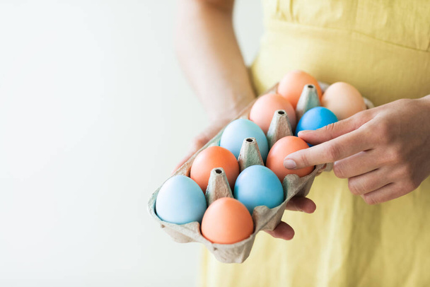 Woman in rustic linen yellow dress holding carton box with painted red and blue eggs for Easter close up. Paschal concept. - Photo, Image